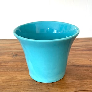 May include: A turquoise ceramic planter with a flared rim. The planter has a smooth, glossy finish and is set on a wooden surface. The planter is empty and ready for plants or flowers.