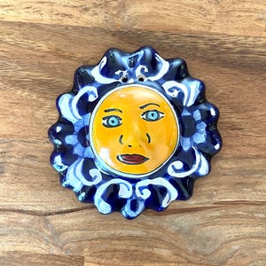 May include: A circular ceramic wall plaque featuring a central yellow sun face with blue eyes and dark red lips. The sun is surrounded by a scalloped border of deep blue glaze with white swirling patterns and two small holes at the top for hanging.