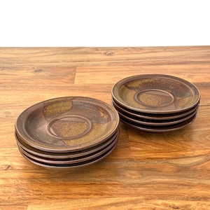 May include: A set of brown and tan ceramic saucers with a circular design. The saucers have a raised center and a dark brown rim. The saucers are stacked in two separate piles on a wooden surface.
