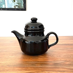 May include: A black ceramic teapot with a lid and a handle. The teapot is sitting on a wooden surface.