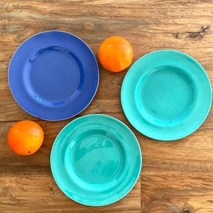 May include: Three colorful ceramic plates in blue and teal, with a gold rim, are arranged on a wooden surface. Two oranges are placed near the plates. The plates are likely for serving food.