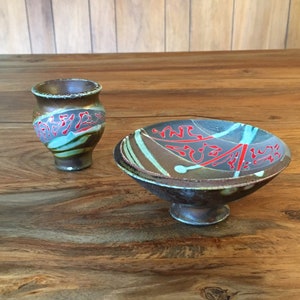 May include: A small, brown ceramic bowl with a white and blue swirl design and red accents sits on a wooden surface. A smaller, brown ceramic cup with a similar design is next to the bowl.