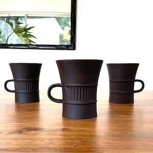 May include: Three dark brown ceramic mugs with handles are arranged on a wooden table. Each mug has a cylindrical band with vertical ridges around the middle. The mugs have a flared top and a small handle.