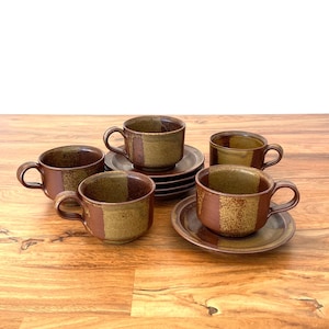 May include: A set of five ceramic teacups and saucers. The cups and saucers are brown and tan with a two-tone design. The cups have handles. The set is arranged on a wooden surface.