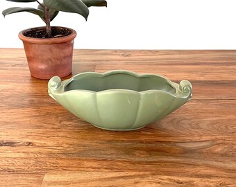 May include: A light green ceramic bowl with a scalloped edge and two handles. The bowl is sitting on a wooden table.