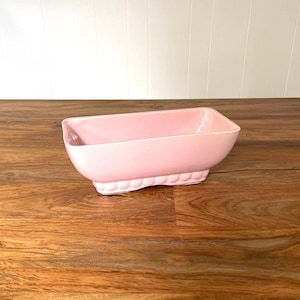 May include: A pale pink ceramic planter with a rectangular shape and a scalloped edge. The planter is sitting on a wooden surface.