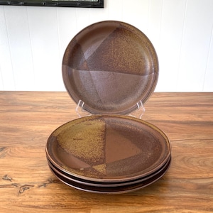 May include: Set of four brown ceramic plates with a geometric design. Each plate features a segmented pattern of dark and light brown tones. The plates are displayed on a wooden surface, with one plate elevated on a clear stand.