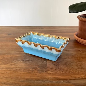 May include: A light blue ceramic dish with a gold rim and a wavy edge. The dish is sitting on a wooden surface.