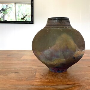 May include: A large, round, dark brown ceramic vase with a textured surface and a black rim. The vase has a subtle iridescent sheen, with hints of purple, blue, and green.