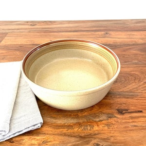May include: A beige ceramic bowl with brown stripes around the rim. The bowl is sitting on a wooden surface.