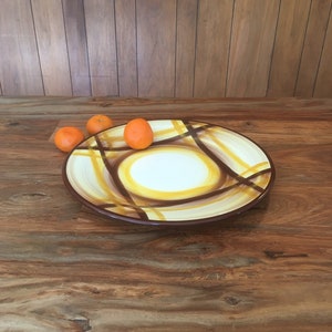 May include: A large, round, ceramic serving platter with a brown rim and a white center. The platter has a yellow and brown geometric design. Three small, orange clementines are sitting on the table next to the platter.