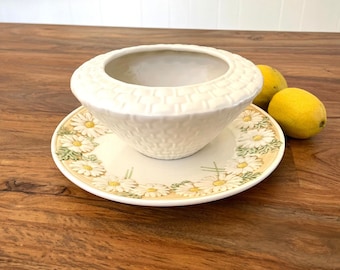 May include: White ceramic bowl with a woven pattern and a matching plate with a daisy design. The bowl and plate are on a wooden surface.