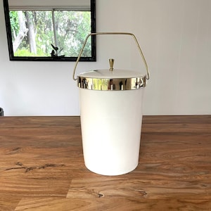 May include: White ice bucket with a gold handle and lid. The bucket is sitting on a wooden surface.