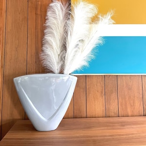 May include: A light gray ceramic vase with a triangular shape, holding a bouquet of white pampas grass. The vase is sitting on a wooden surface in front of a wall with a blue, white, and yellow striped pattern.