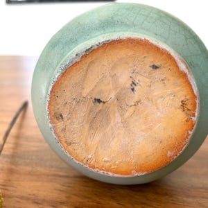 May include: A close-up of the bottom of a light green ceramic vase. The base is a light brown color with a rough texture and a darker brown ring around the edge. The vase is sitting on a wooden surface.