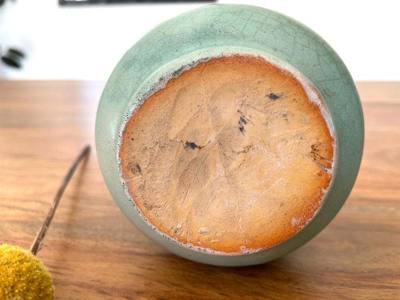 May include: A close-up of the bottom of a light green ceramic vase. The base is a light brown color with a rough texture and a darker brown ring around the edge. The vase is sitting on a wooden surface.
