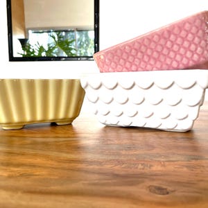 May include: Three ceramic bowls in a stack on a wooden table. The top bowl is pink with a diamond pattern. The middle bowl is white with a scalloped edge. The bottom bowl is yellow with a ribbed design.