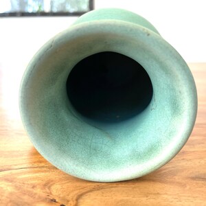 May include: A close-up view of a light green vase with a wide, flared opening. The vase has a smooth, matte finish and a dark interior. The vase is sitting on a wooden surface.
