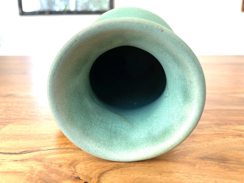 May include: A close-up view of a light green vase with a wide, flared opening. The vase has a smooth, matte finish and a dark interior. The vase is sitting on a wooden surface.