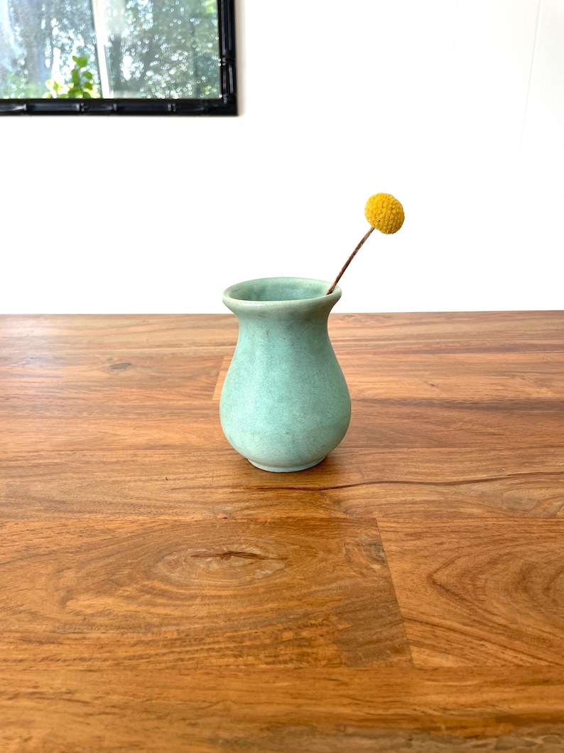 May include: A small, light-green ceramic vase with a rounded body and flared rim. A single yellow flower with a round head is placed in the vase. The vase is on a wooden surface.