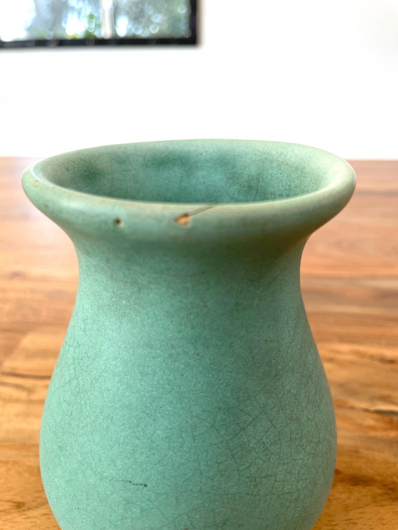 May include: A celadon green ceramic vase with a flared rim and a textured surface. The vase has a crackled appearance and a small chip on the rim. The vase is a decorative item.