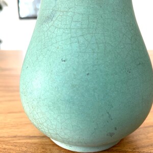 May include: A light teal-colored ceramic vase with a crackle glaze finish. The vase has a rounded body and a narrow neck, sitting on a wooden surface. The surface of the vase has a network of fine cracks.