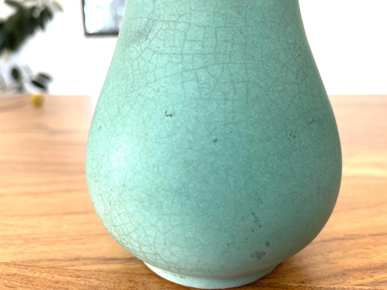 May include: A light teal-colored ceramic vase with a crackle glaze finish. The vase has a rounded body and a narrow neck, sitting on a wooden surface. The surface of the vase has a network of fine cracks.