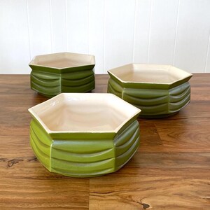 May include: Three green ceramic planters with a ribbed design. The planters have a hexagonal shape and a cream-colored interior. They are arranged on a wooden surface.