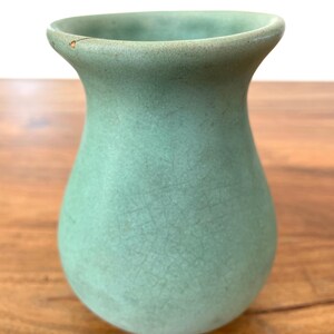 May include: A small, celadon green vase with a crackled texture. The vase has a rounded body and a flared rim. The vase is displayed on a wooden surface.