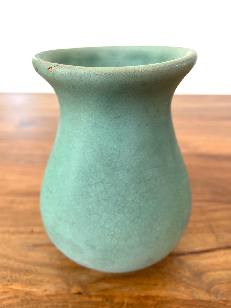May include: A small, celadon green vase with a crackled texture. The vase has a rounded body and a flared rim. The vase is displayed on a wooden surface.