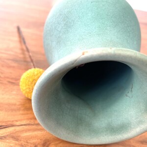 May include: A light green ceramic vase with a wide opening and a textured surface. A small yellow pom-pom on a brown stem rests beside the vase. The vase is on a wooden surface.
