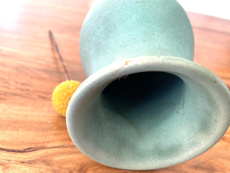 May include: A light green ceramic vase with a wide opening and a textured surface. A small yellow pom-pom on a brown stem rests beside the vase. The vase is on a wooden surface.