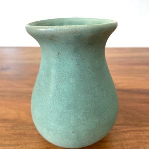 May include: A small, celadon-colored ceramic vase with a crackle glaze. The vase has a rounded body and a flared rim. The color is a muted green-blue, and the surface has a textured appearance. The vase is likely for decorative purposes.