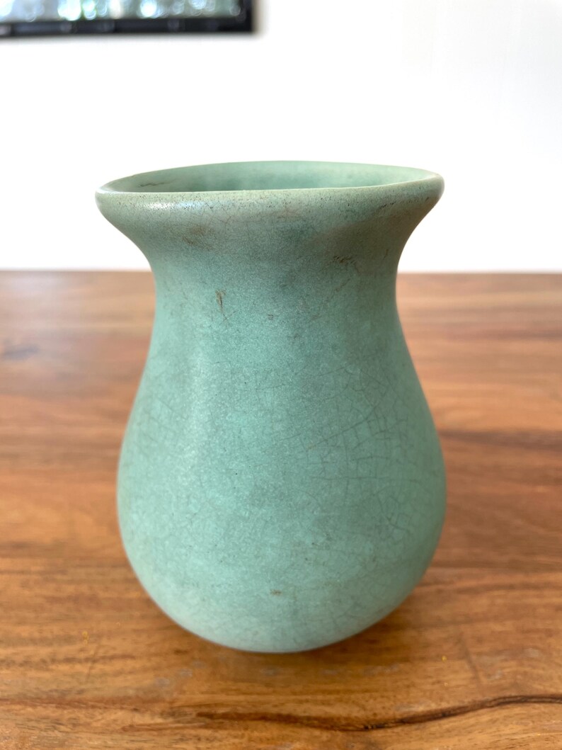 May include: A small, celadon-colored ceramic vase with a crackle glaze. The vase has a rounded body and a flared rim. The color is a muted green-blue, and the surface has a textured appearance. The vase is likely for decorative purposes.