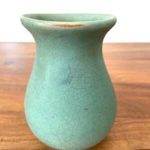 May include: A small, light green ceramic vase with a crackle glaze. The vase has a rounded body and a flared rim. The color is a muted, seafoam green with subtle variations in tone. The vase is sitting on a wooden surface.