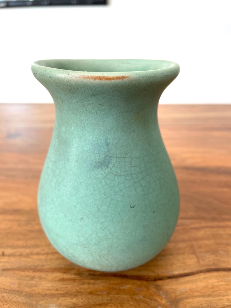 May include: A small, light green ceramic vase with a crackle glaze. The vase has a rounded body and a flared rim. The color is a muted, seafoam green with subtle variations in tone. The vase is sitting on a wooden surface.