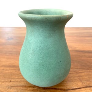 May include: A small, light green ceramic vase with a rounded body and flared rim. The vase has a textured surface and sits on a wooden table. The vase is a decorative item.