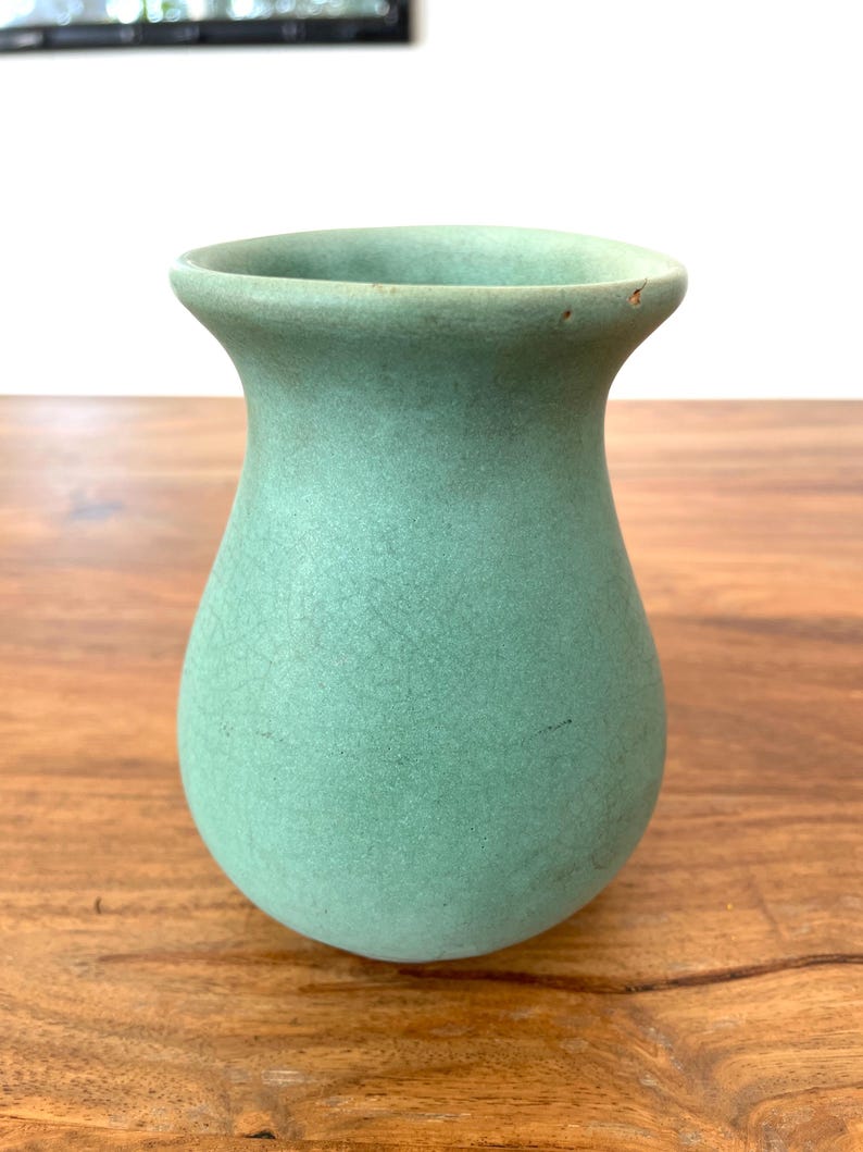 May include: A small, light green ceramic vase with a rounded body and flared rim. The vase has a textured surface and sits on a wooden table. The vase is a decorative item.