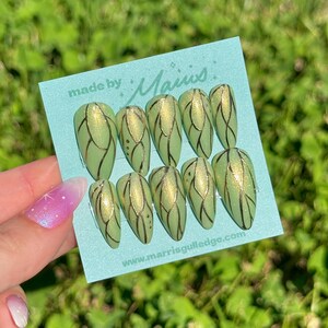 May include: A set of press-on nails in a light green color with gold and black leaf-like designs. The nails are displayed on a light blue card with the text "made by Maius" and a website address.