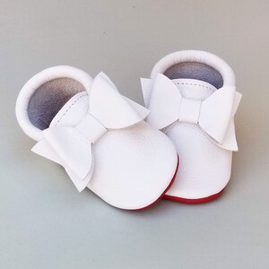 Puede incluir: Un par de mocasines blancos para beb&eacute; con grandes lazos decorativos. Los zapatos tienen suelas rojas y una textura suave similar al cuero. El interior es de color gris claro y los zapatos est&aacute;n dise&ntilde;ados para beb&eacute;s.