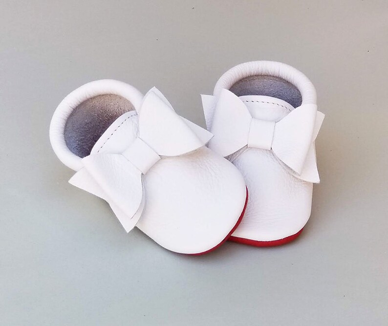 Puede incluir: Un par de mocasines blancos para beb&eacute; con grandes lazos decorativos. Los zapatos tienen suelas rojas y una textura suave similar al cuero. El interior es de color gris claro y los zapatos est&aacute;n dise&ntilde;ados para beb&eacute;s.