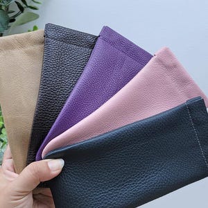 Puede incluir: Una colección de estuches de cuero para gafas en varios colores, incluyendo beige, negro, morado, rosa y azul marino. Los estuches son de cuero texturizado y están dispuestos en forma de abanico.
