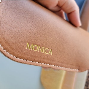 May include: A brown leather glasses case with a white stitch around the edge. The case has the name "MONICA" embossed in gold lettering.