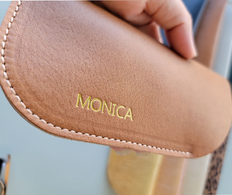 May include: A brown leather glasses case with a white stitch around the edge. The case has the name "MONICA" embossed in gold lettering.