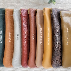 May include: A collection of leather pouches in various colors, including Green Forest, Pumpkin, White, Mahogany, Brick, Antique Camel, Honey Mustard, Distressed Espresso, and Mustard Green. Each pouch has a gold-colored metal clasp and the color name printed on it.