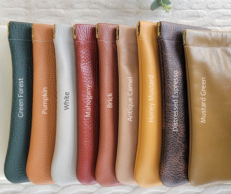 May include: A collection of leather pouches in various colors, including Green Forest, Pumpkin, White, Mahogany, Brick, Antique Camel, Honey Mustard, Distressed Espresso, and Mustard Green. Each pouch has a gold-colored metal clasp and the color name printed on it.