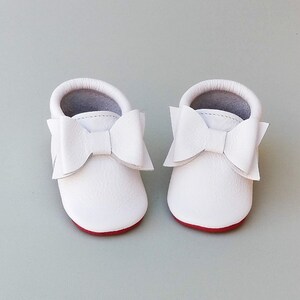 Puede incluir: Un par de mocasines blancos de cuero para beb&eacute; con grandes lazos decorativos. Los zapatos tienen suelas rojas e interiores grises. Estos zapatos de beb&eacute; de suela blanda est&aacute;n dise&ntilde;ados para la comodidad y el estilo.