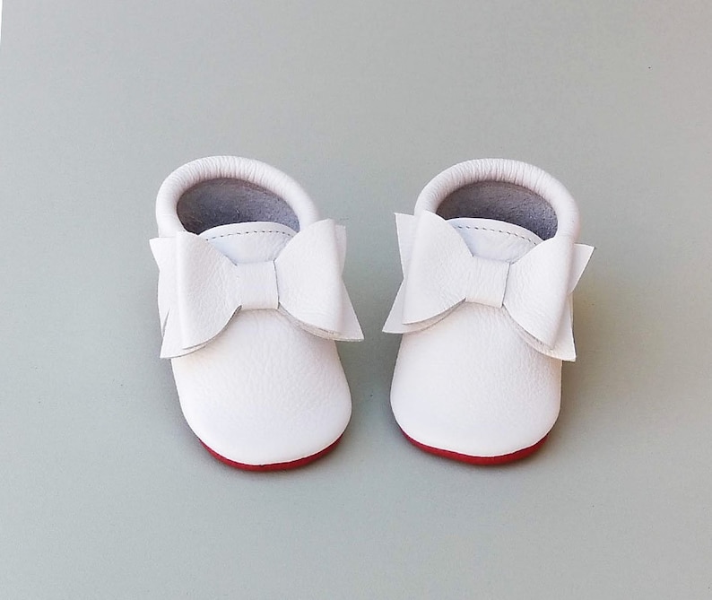 Puede incluir: Un par de mocasines blancos de cuero para beb&eacute; con grandes lazos decorativos. Los zapatos tienen suelas rojas e interiores grises. Estos zapatos de beb&eacute; de suela blanda est&aacute;n dise&ntilde;ados para la comodidad y el estilo.