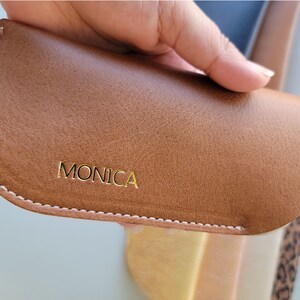 May include: A brown leather glasses case with white stitching and the word "MONICA" embossed in gold lettering.