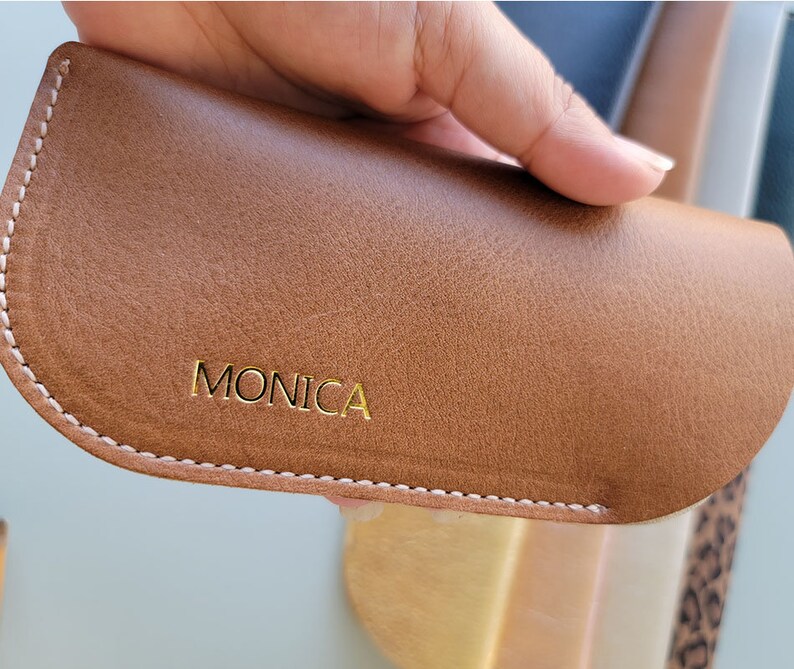 May include: A brown leather glasses case with white stitching and the word "MONICA" embossed in gold lettering.